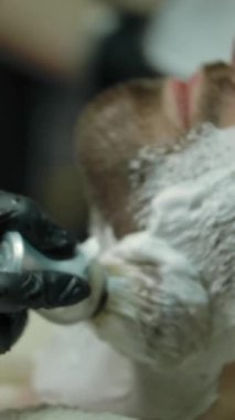 Modern berber dükkanında tıraş olmadan önce insanın yanağına ve boynuna köpük sürmek için fırça kullanan eldivenli berber. Dikey çekim.