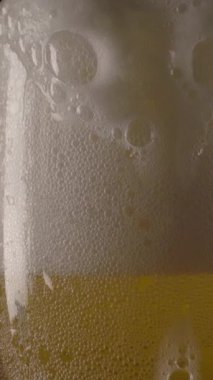 Koyu bardaki cam bardağın kenarından dökülen yumuşak soğuk bira köpüğü. Dikey çekim.