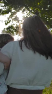 Kucağında kızı ile beyaz elbiseli anne yeşil bir alanın ortasında bir salıncak ile eski bir meşe ağacına gider. Yavaş çekim. Güneş parıltısı yaprakları ile güneşin parlama yapar. Geri görünüm