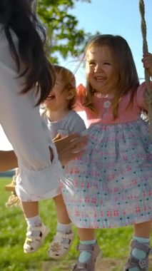 Beyaz elbiseli mutlu esmer kadın yeşil bir tarlanın ortasında kızlarını salıncakta sallıyor. İki bebek gülüyor ve gülümsüyor. BGG 'de ağaç dalı ve mavi gökyüzü. Ağır çekim. Dikey