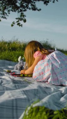 Üç yaşındaki bir kız çayırda bir çay partisinde yumuşak oyuncaklarıyla oynuyor. Annem salıncakta sallanıyor, atlar ve bir yaşındaki kız kardeşine yardım etmek için koşar. Yavaş çekim gimbal stabilize atış. Gün batımı doğal