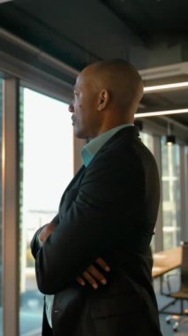 Ciddi bir ifadeyle modern ofis penceresinin yanında duran Aframerikalı girişimci. İş alanı geçmişi. 4k UHD süzülme izi portresi. Dikey çekim.