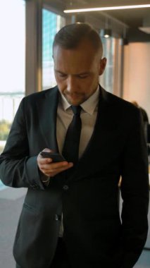 Pencerenin yanında duran ve SMS yazan çekici bir iş adamının portresi. Çok zeki, ciddi ve ağır çekimde görünüyor. Kaydırma kamerası hareketi. Dikey çekim.