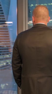 Takım elbiseli başarılı bir işadamının elleri ceplerinde ofiste yürümesi. Arka taraftaki işadamı pencerenin önündeki şehir manzarasının önünde. Orta 4k Ultra HD çekimi. Dikey çekim