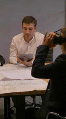 Sanal gerçeklik gözlüklü kendine güvenen girişimci. Modern cam ofiste inşaat ve yol çizimleri üzerinde çalışan bir grup genç müteahhit. Çeşitli iş adamları toplantı odasını arıyor. Dikey çekim.