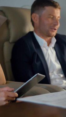 Kendine güvenen zengin meslektaşlar birinci sınıf uçuş sırasında iş konuşuyorlar. Tablet bilgisayara ve belgelere odaklanıyorum. Parlak gün ışığı lombozu. Dikey çekim.