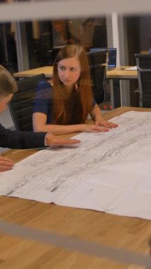 Masadaki meslektaşlar çizim kağıdına bakıp konuşuyorlar. Resmi ofis kıyafeti giymiş bir personel masanın arkasında oturup tartışıyor. Geniş 4k UltraHD yavaş çekim. Cam manzarayı fırlat. Dikey