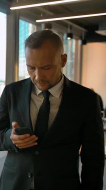 Resmi takım elbiseli çekici girişimci pencerenin yanında duruyor ve mesaj yazıyor. Çok ciddi ve zeki görünüyor. Ağır çekim. Dikey çekim.