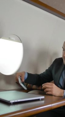 Özel jetiyle seyahat eden ve cep telefonuyla sohbet eden resmi takım elbiseli kendine güvenen bir iş adamı. Pahalı Macbook 'u kapattı ve iPhone' unu elinde tuttu. Dikey çekim.