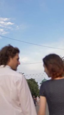 Genç bir çift bir Avrupa kasabasında yaya caddesi boyunca yürür. El ele tutuşur, gülümser, güler ve konuşurlar. Sıcak yaz akşamı, gün batımı doğal ışık. Arka orta görünüm dikey çekim.