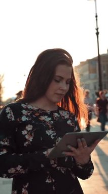 Şehir sokaklarında gün batımında tabletli genç çekici kızıl saçlı kız internette sörf yapıyor. İlginç bir şey bulmaya çalışıyor. Arka planda bir sürü insan var. Yavaş çekim dikey çekim.