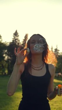 Genç bir kadın sabun köpüğüyle oynuyor, dışarıda eğleniyor. Yeşil yaz akşamı günbatımı parkında yürüyor. Süper yavaş çekim. Dikey çekim.