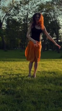 Parkta zıplayan ve dans eden yakışıklı genç kız. O, zaman geçirmekten mutlu. Gün doğumu ya da batımı güzel bir ışık. Gerçek zamanlı ve yavaş çekim dikey çekim.