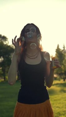 Genç bir kadın sabun köpüğüyle oynuyor, dışarıda eğleniyor. Yeşil yaz akşamı günbatımı parkında yürüyor. Süper yavaş çekim. Dikey çekim.