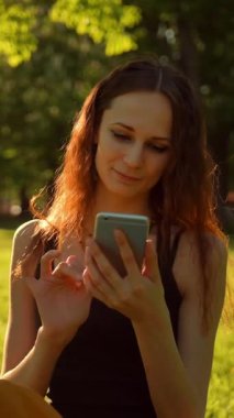 Mutlu Gülümseyen Genç Kadın doğanın ve güneşin tadını çıkarıyor. Yeşil yaz parkında cep telefonuyla bankta oturmak. Neşeli bir bayan cep telefonuyla konuşuyor, akıllı telefondan arkadaşlarıyla sohbet ediyor. Orta çekim
