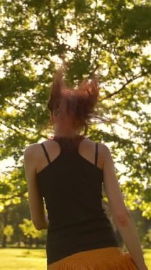 Uzun dalgalı kızıl saçlı genç bayan ormanda koşuyor ve zıplıyor yaz mevsiminde güneşli akşam günbatımında, kameraya dönüyor. Günbatımı veya gündoğumu Dikey çekim.