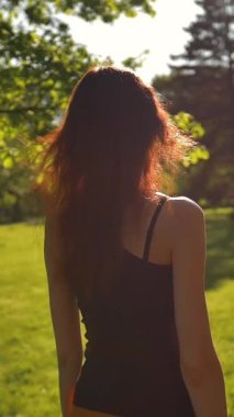 Güzel, çekici, seksi, kızıl saçlı bayan parkta temiz havanın ve doğanın tadını çıkarıyor, sonra dönüp gülüyor. Süper yavaş çekim. Gün batımı. Dikey çekim.