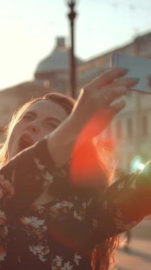 Sokakta çekilmiş, modaya uygun genç bir kadının şehirde selfie çekerken çekilmiş bir görüntüsü. Öpüşme ve saçlarıyla oynadığını hissediyor. Ağır çekim. Dikey çekim.