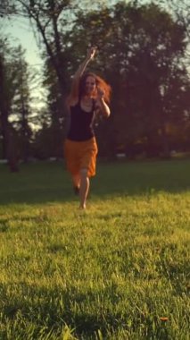 Gün batımında parkta çimenlerde dans edip zıplayan genç bir kadın. Mutlu kadın dans ediyor. Orta plan. Sıcak bir yaz günü. Süper yavaş çekim. Dikey çekim.