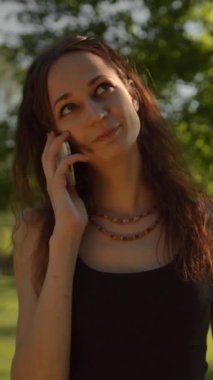 Parkta durup telefonla konuşan gülümseyen genç bayan mutlu ve neşeli görünüyor. 4K ağır çekim. Dikey çekim.