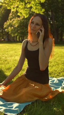 Parktaki bir telefon konuşması sırasında kameraya doğru oturan bir kadının ön görüntüsü. Yavaş çekim. Dikey çekim.