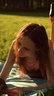 Parkta çimlerin üzerinde oturan yakışıklı seksi kadın. Kablosuz internet bağlantısı. Sosyal ağ kurmak ve mobil iletişim için sohbet etmek. Dijital ped veya tabletPC ortadaki 4k UHD dikey çekim.