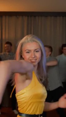 Partide dans eden kadın. Sarı elbiseli neşeli sarışın kız arkadaşının doğum gününde dans ediyor. Arkadaşları arka plandaki havalı müzik ölçüsüyle mutlu. Ağır çekim. 4k dikey çekim.