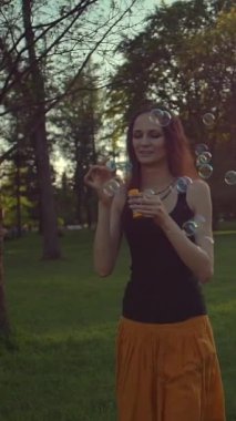 Genç bir kadın sabun köpüğüyle oynuyor, dışarıda eğleniyor. Güneş parlaması. Yeşil yaz sabahı gündoğumu parkında yürüyor. Süper yavaş çekim orta çekim. Dikey çekim.