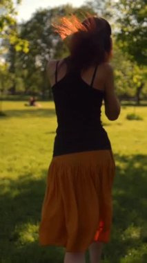 Uzun saçlı güzel bir kızıl kadın. Kulaklıklı müzikten hoşlanıyor. Parkta koşuyor. Çocukluktaki gibi zıplıyor. Dikey çekim.