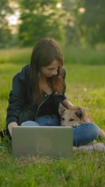 Açık bir dizüstü bilgisayarın yanındaki parkta bir kız köpeğini okşuyor. İçeriği dışarıda dizüstü bilgisayar kullanarak tüketmek. Parkta yürüyüş için favori evcil hayvan. Dikey çekim.