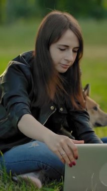Serbest çalışan bir kız köpeğinin yanındaki yeşil bir çayırda otururken iş için bilgisayarını açıyor. Akşamları dışarıda çalışma zamanıdır. Yaz gün batımı ışığı. Dikey çekim.