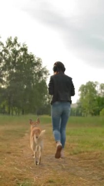 Kulaklıklı çekici bir kadın parkta köpeğiyle yürüyor. Kot pantolonlu ve siyah deri ceketli bir kadın köpeğiyle yolda yürüyor. Kadın müzik dinliyor. Arka plan, yavaş çekim