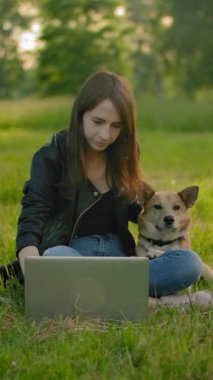 Parkta dizüstü bilgisayarın yanında güzel beyaz bir kadın köpeğini okşuyor. İçeriği dışarıda dizüstü bilgisayar kullanarak tüketmek. Favori köpeğim sakince parkta oturuyor. Orta atış. Dikey