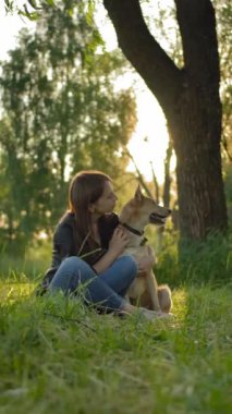 Hanımefendiyle köpeği arasındaki hassas ilişki. Deri ceketli ve mavi kotlu bir kızın köpeğiyle geniş açılı görüntüsü. Gün batımında uzun bir ağacın yanındaki çimlerin üzerinde oturuyorlar.