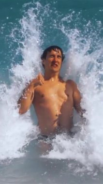 Ağır çekim. Denizde bel hizasında duran genç adam, Atlantik Okyanusu 'nun köpük dalgası tarafından yere serilen baş parmağını kaldırıyor. Teal ve turuncu dikey çekim.