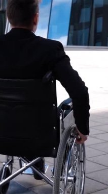 Tekerlekli sandalyedeki genç adam modern cam iş merkezinin yakınına taşınıyor. Arkadan bak. Süper yavaş çekim. Kötü negatif duygu dikey çekim.