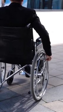 Tekerlekli sandalyedeki genç iş adamı modern cam iş merkezinin yakınına taşınıyor. Arkadan bak. Süper yavaş çekim. Olumsuz duygu, başını çeviriyor. Resmi takım elbise. Dikey çekim.