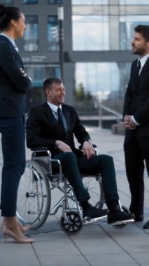 Tekerlekli sandalyedeki özürlü beyaz adam ve iş arkadaşları günbatımında modern ofis binasının yakınında sohbet ediyorlar. Dikey çekim.