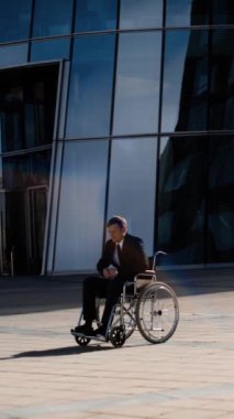 Tekerlekli sandalyedeki üzgün, yakışıklı, engelli girişimci işadamı iş bölgesinde ve modern yapım turuncusunda ilgisiz bir kalabalık. Dikey çekim.