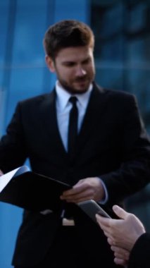 Engelli iş arkadaşlarıyla dışarıda iş konuşuyorlar. Tekerlekli sandalyede oturan ve ipad, not defteri ve belgelerle çalışan bir adam. Modern iş merkezi binası bg. Orta atış. Dikey