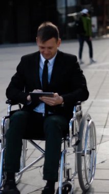 Genç gülümseyen engelli işadamı dışarıda tekerlekli sandalyede oturuyor ve tabletPC açık hava ve modern parlak binalarla çalışıyor. Dikey çekim.