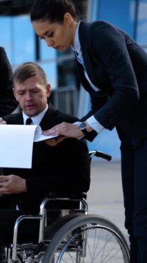 Tekerlekli sandalyedeki iş adamı iş ve anlaşma hakkında konuşuyor. Metin defteri, tablet bilgisayar, cep telefonu, cep telefonu ve belgelerle çalışıyorlar. Toplumun bir parçası olan topal insanlar. Dikey çekim.