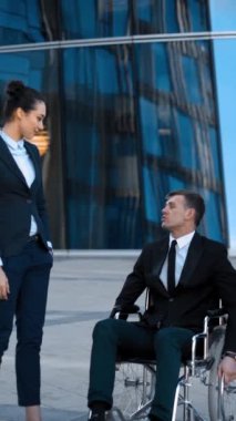 Tekerlekli sandalyedeki engelli girişimci işadamı ve meslektaşları dışarıda gülüp eğlenerek olumlu bir konuşma yapıyorlar. Kameraya bakarlar. 4k... Teal ve turuncu atış. Dikey çekim.
