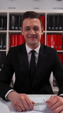 4k UHD. Genç başarılı pazarlamacı girişimci işadamı masası, cep telefonu ve dijital bloknotu olan modern ofis içindeki komik şakanızla işaret ediyor ve gülüyor. Dikey Çekim.