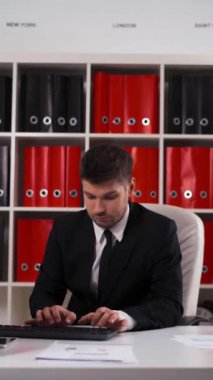 4k UHD. Siyah takım elbiseli ve kravatlı başarılı genç ve çekici iş adamı ofiste bilgisayarla çalışıyor. Beyaz, siyah ve kırmızı yüzgeçli tabletli, cep telefonlu. Kameraya gülümsüyor. Dikey