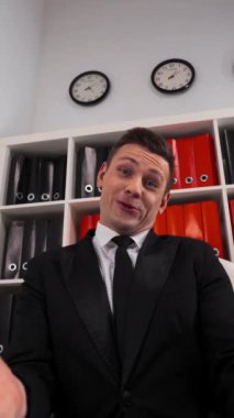 Young attractive businessman in a black suit and tie sitting inside clean white office showing gesture fuck joke and smile. Middle finger to You POV. UltraHD 4k Vertical Shot.