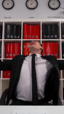 Resmi takım elbiseli ve kravatlı genç ve çekici bir iş adamı ofisin içinde oturmuş dans ediyor. 4K UHD ile geniş açı. Dikey Çekim.