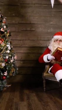 Noel Baba, Home 'daki Noel Ağacı yakınlarındaki kitap ya da fotoğraf albümünü çekip okuyor. İçinde hediyeler ve şömine olan bir çanta. Geniş aç. Dikey Çekim.