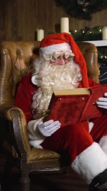 Noel Baba Noel arifesinde koltukta oturmuş yeşil yılbaşı ağacının yanında üzerinde oyuncaklar ve ışıklar olan eski kırmızı ipek albümleri çıkarıyor. Geniş açılı kaydırma doli hareketi Dikey Çekim.