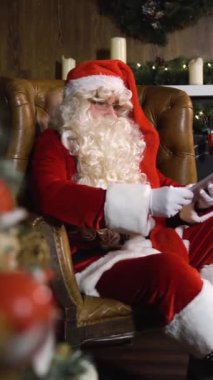 Noel Baba tablet PC IPad ile çalışıyor. Işıkları ve arka planda süslü şöminesi olan Noel ağacı..